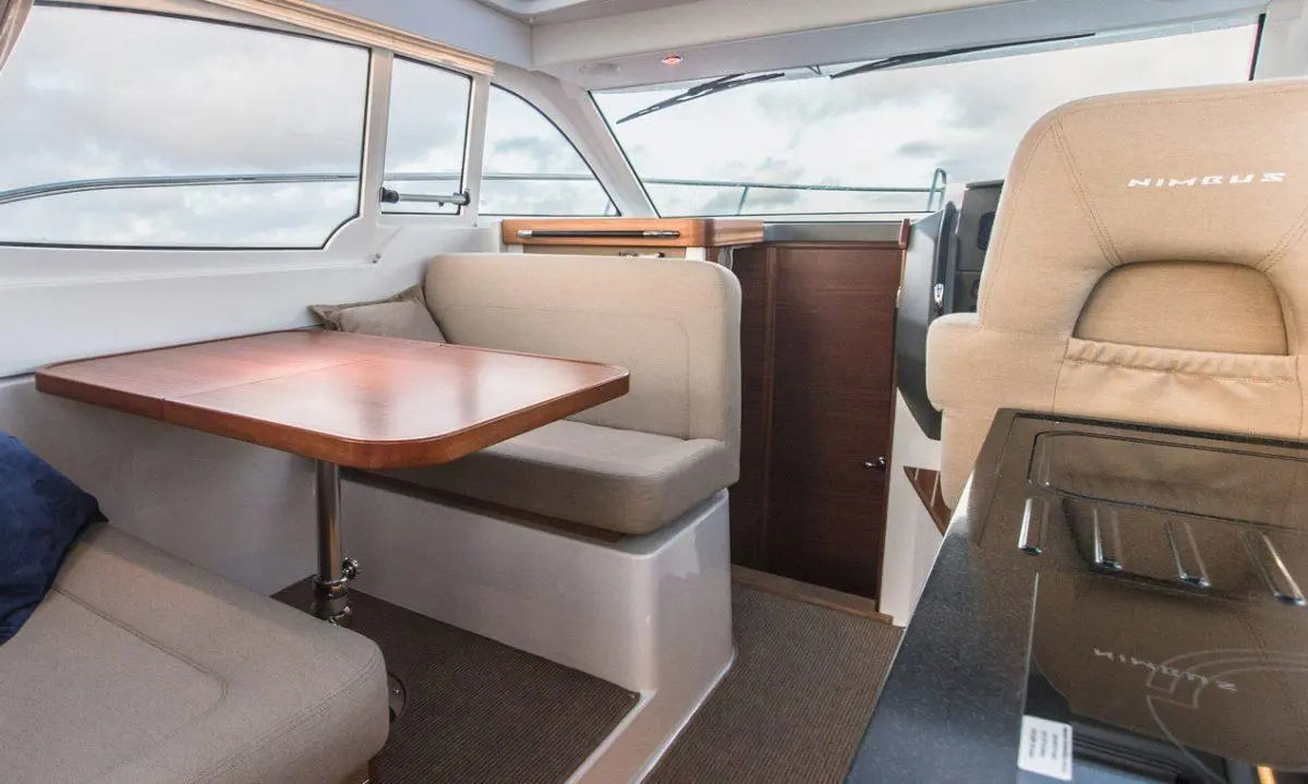 Interior of a Nimbus boat showing a wooden dining table, seating area, and helm with large windows and light upholstery.